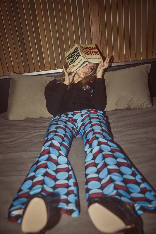 Woman sitting on a bed holding an open book over her face, wearing patterned blue flared trousers and a dark top, photographed by Amanda Pratt.