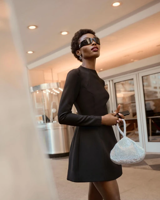Stylish woman in a black dress and sunglasses holds a beaded bag in an urban setting, creating a fashion-forward and chic aesthetic.
