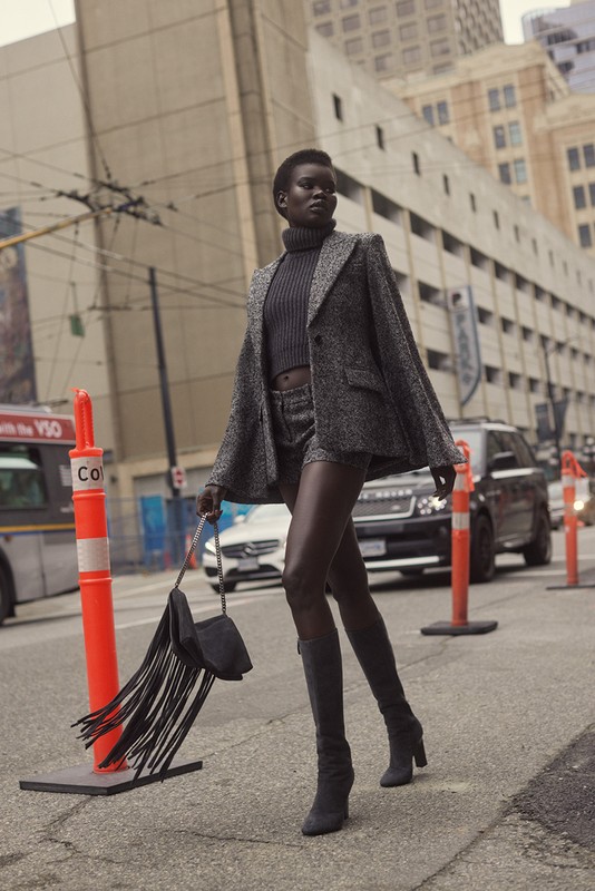 Fashion editorial shot of a Black woman walking in a tweed jacket and shorts on a city street with buildings in the background.