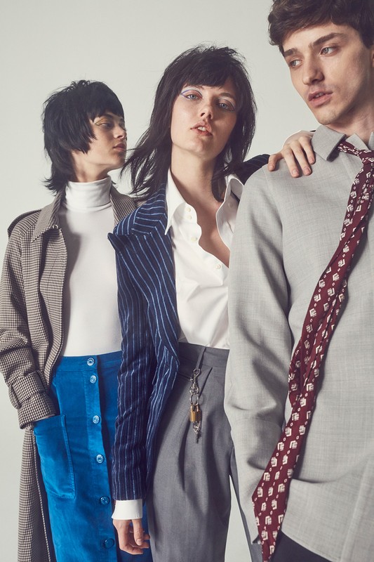 Three fashion models pose in a studio, showcasing a pinstripe blazer, trench coat, collared shirt, and a patterned tie in a neutral setting.