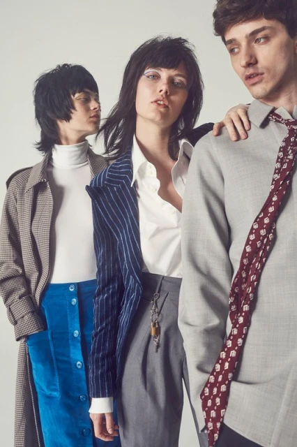 Three fashion models pose in a studio, showcasing a pinstripe blazer, trench coat, collared shirt, and a patterned tie in a neutral setting.