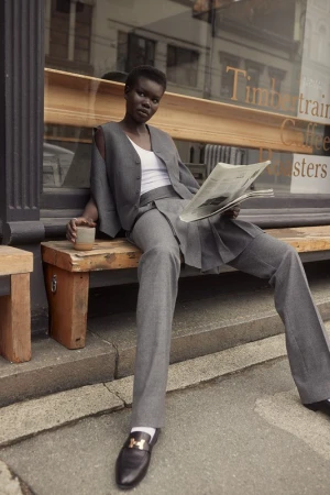 Black woman wearing grey suit trousers and vest holding a newspaper while sitting on a wooden bench outdoors