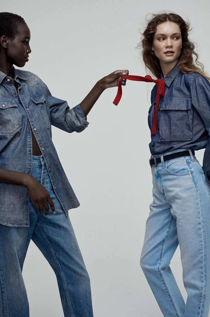 Two women in denim outfits pose in a studio, one with dark skin, the other with light skin and a red scarf. Fashion shot.