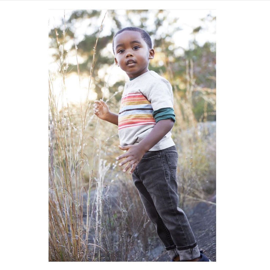 A young, black child stands in a field of tall grass wearing a striped shirt and dark jeans in an outdoor setting during the daytime.