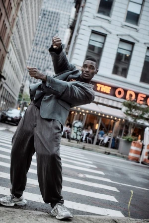 Man wearing a grey suit and sneakers dancing on a city crosswalk with a restaurant storefront and tall buildings in the background.