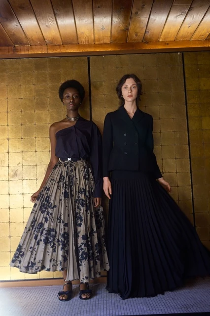 Two women in elegant vintage-inspired fashion: one in a one-shoulder blouse and floral skirt, the other in a black suit and pleated skirt.