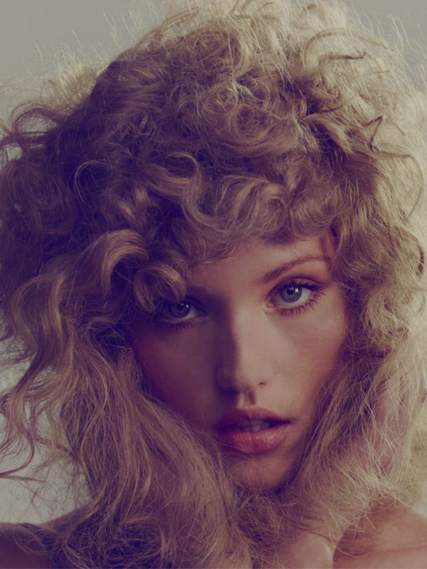 Close-up of a woman with curly blonde hair, fair skin, and blue eyes, posing in a studio with soft, diffused lighting on neutral background.