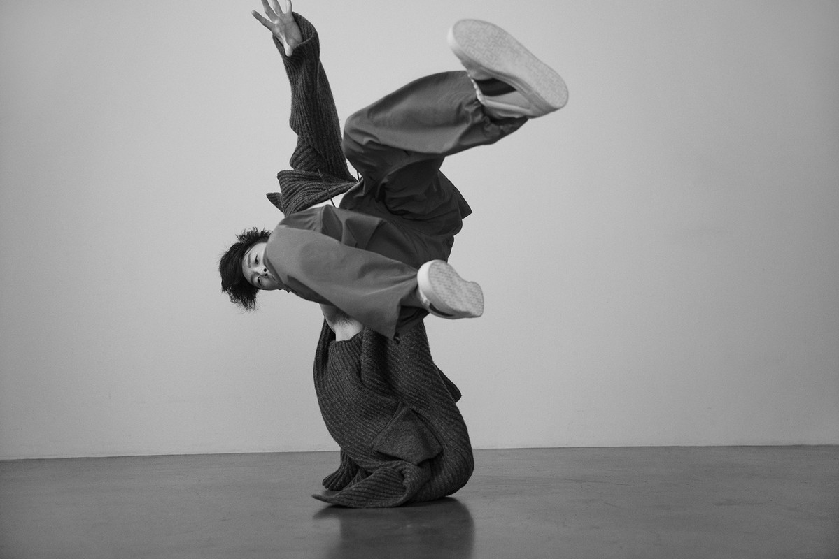 Monochrome portrait of a non-binary person in a handstand wearing sweater and pants on a grey background in a photo studio.