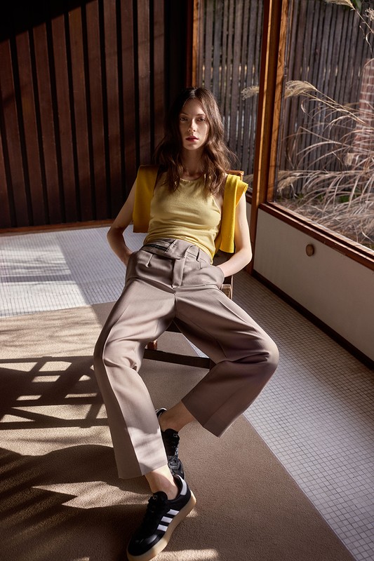 A woman in a yellow top and taupe pants sits indoors near a window, with natural light casting shadows across the floor. She wears black sneakers.