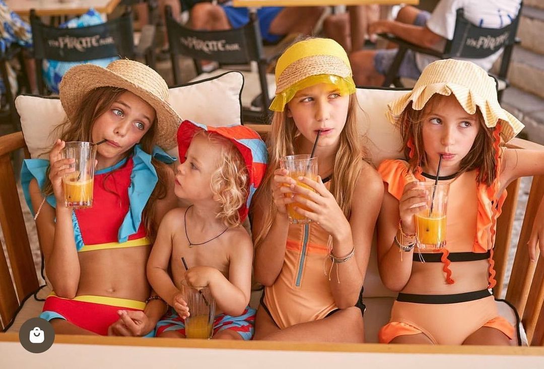 Group of four children in swimsuits seated, holding juice glasses, wearing summer hats. Beach chairs and sunlit outdoor setting.