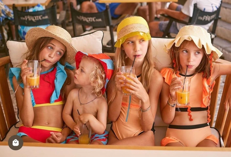 Group of four children in swimsuits seated, holding juice glasses, wearing summer hats. Beach chairs and sunlit outdoor setting.