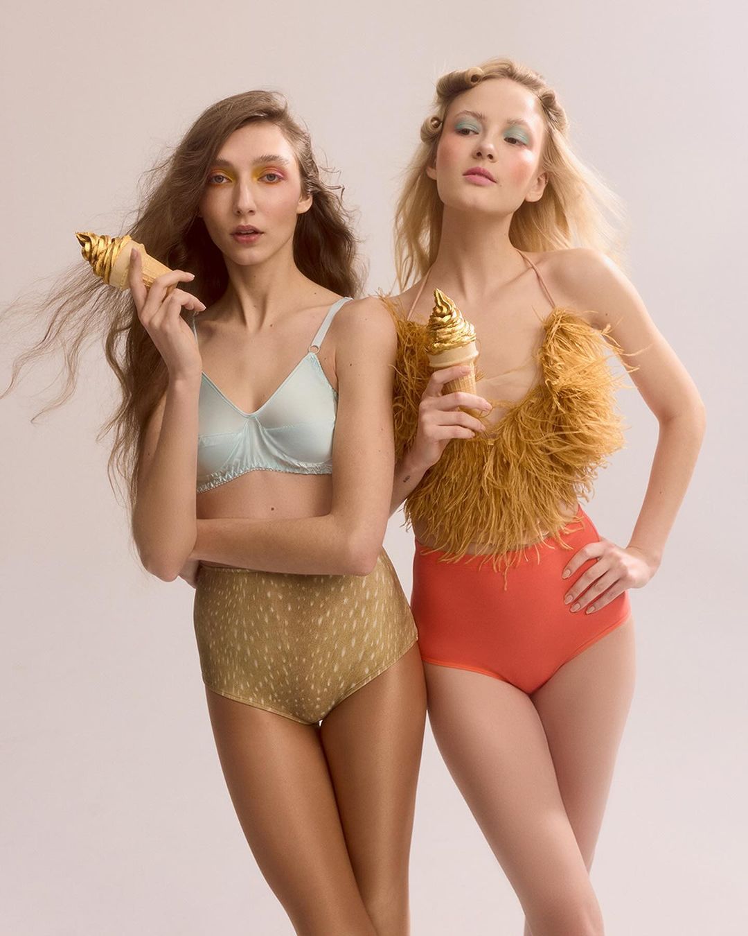 Two women in fashionable vintage swimwear holding golden ice cream cones, standing in front of a plain backdrop in a studio setting.