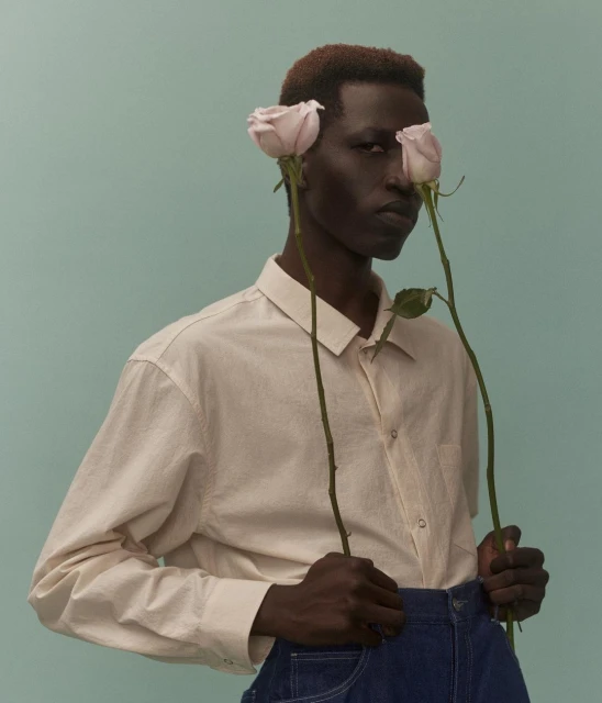 Portrait of a young adult man with dark skin and short hair, wearing a light button-down shirt and holding two pink roses against a blue background.