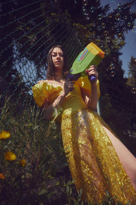 A woman in a yellow dress holds a bubble gun surrounded by trees and greenery, creating a whimsical and playful outdoor scene.