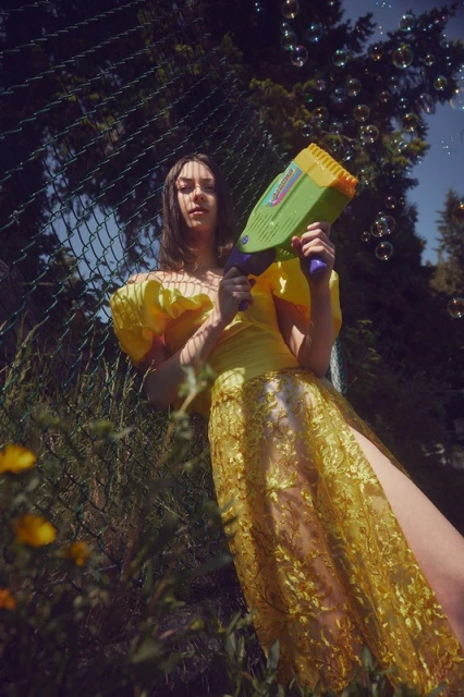A woman in a yellow dress holds a bubble gun surrounded by trees and greenery, creating a whimsical and playful outdoor scene.