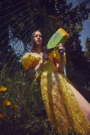 A woman in a yellow dress holds a bubble gun surrounded by trees and greenery, creating a whimsical and playful outdoor scene.