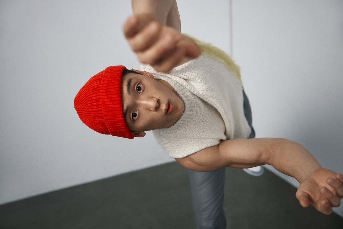 Portrait of an adult Asian male wearing a red beanie and cream-colored sweater, shot from a low angle against a white wall and gray floor.