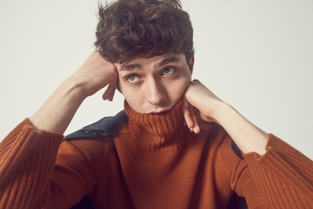Close-up of a young adult male with curly brown hair wearing an orange turtleneck sweater in a brightly lit studio setting.