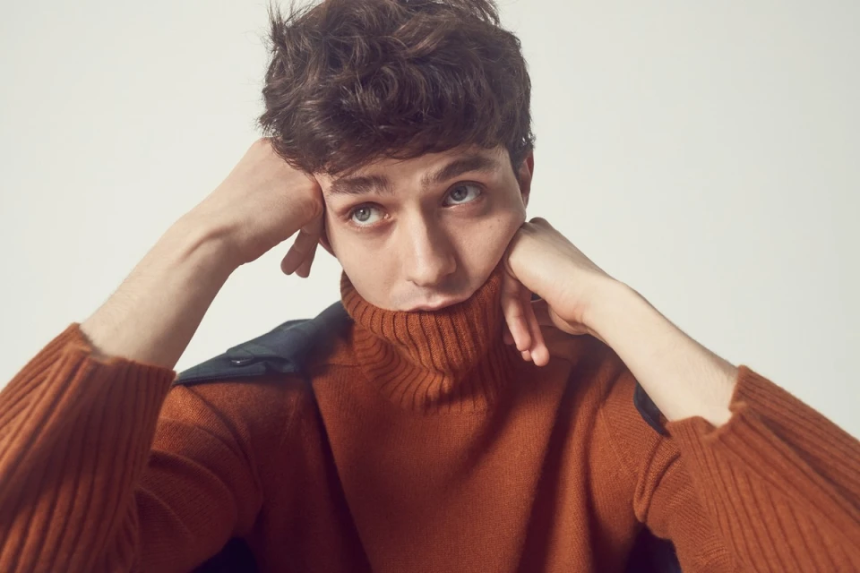 Close-up of a young adult male with curly brown hair wearing an orange turtleneck sweater in a brightly lit studio setting.