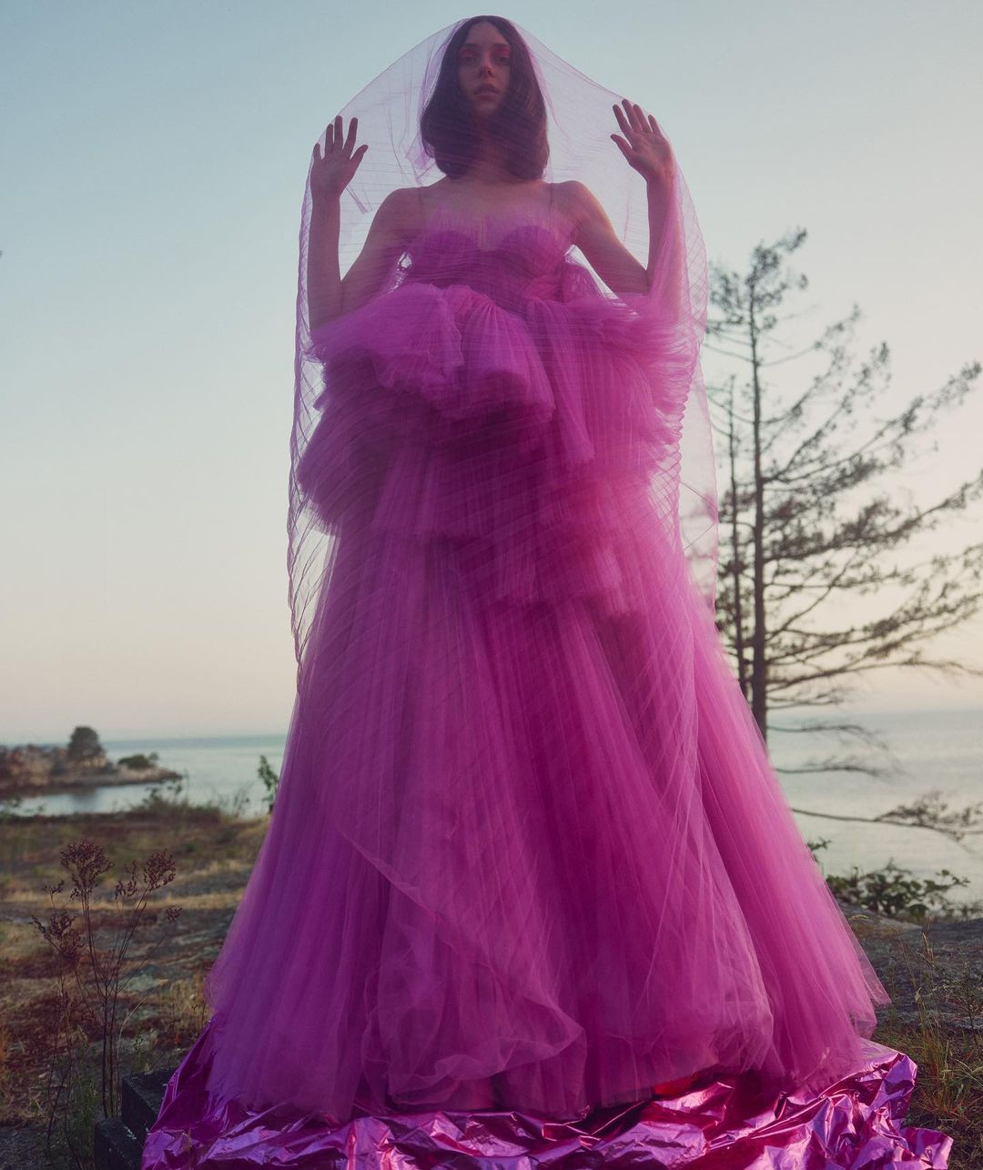A woman in a vibrant pink strapless tulle gown with a long, sheer veil stands on a rocky landscape near the ocean under a bright sky.