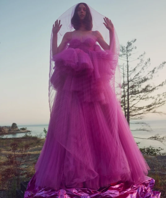 A woman in a vibrant pink strapless tulle gown with a long, sheer veil stands on a rocky landscape near the ocean under a bright sky.