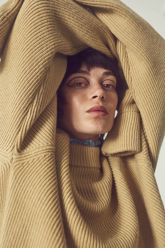 Close-up studio shot of a fair-skinned woman with bangs wearing a tan turtleneck sweater, hands above head, and a blue ruffled collar.
