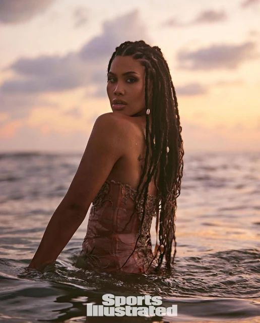 Woman with long braids poses in the ocean at sunset, wearing a pink bodice, as waves lap around her waist and Sports Illustrated logo visible.