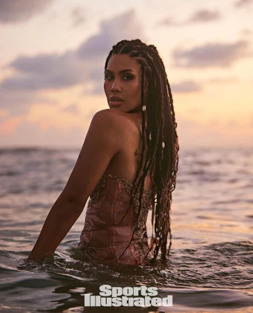 Woman with long braids poses in the ocean at sunset, wearing a pink bodice, as waves lap around her waist and Sports Illustrated logo visible.
