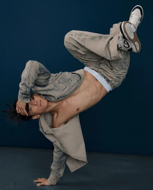 An athletic Asian man breakdancing in a studio wearing a neutral denim jacket and pants with white sneakers, captured in a dynamic pose.