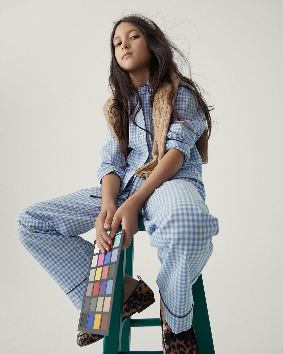 A young girl in a blue-and-white gingham pajama set sits on a green stool, holding a color palette in a studio setting with soft lighting.