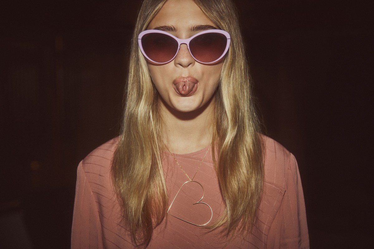 A young woman wearing pink sunglasses and a pink shirt sticks her tongue out, accessorized with a large heart pendant necklace.