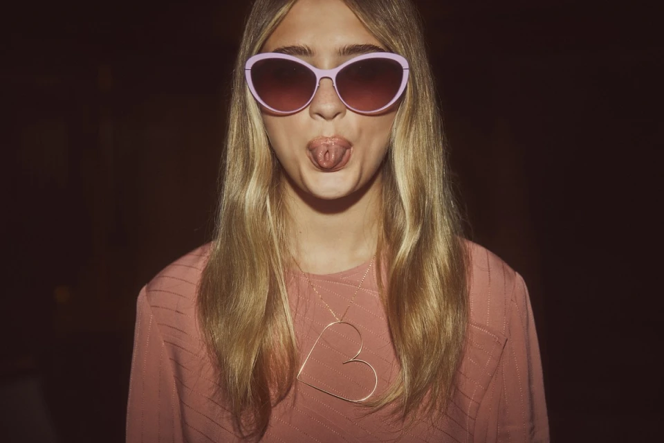 A young woman wearing pink sunglasses and a pink shirt sticks her tongue out, accessorized with a large heart pendant necklace.