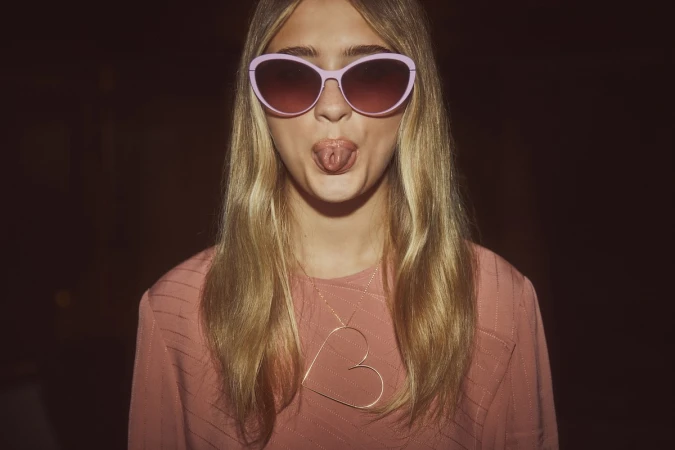 A young woman wearing pink sunglasses and a pink shirt sticks her tongue out, accessorized with a large heart pendant necklace.