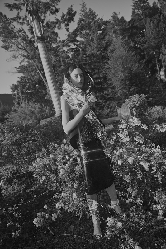 Black-and-white portrait of an adult female model in a black dress and floral scarf, posing outdoors among roses near a column.