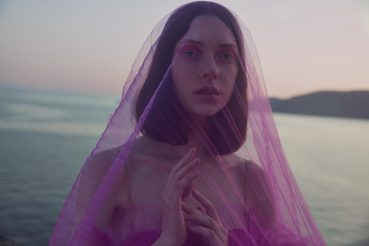 Close-up of a young adult woman with short brown hair and bright red makeup, draped in translucent purple fabric against a waterscape.
