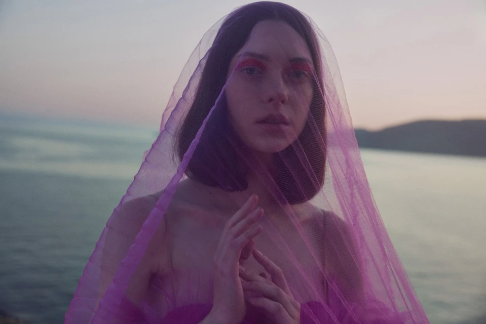 Close-up of a young adult woman with short brown hair and bright red makeup, draped in translucent purple fabric against a waterscape.
