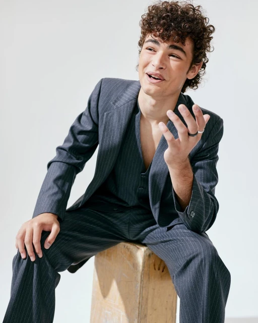Young man with curly hair poses in a gray pinstripe suit sitting on a wooden block against a white backdrop in a studio setting.