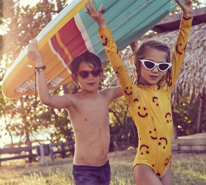 Two kids in swimwear, one holding a surfboard. A boy with red sunglasses, shirtless next to a girl in a yellow smiley-face swimsuit.
