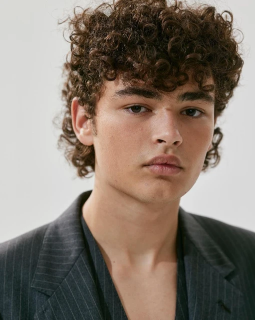 Headshot of a young adult male with curly hair, wearing a dark pinstripe blazer in a studio setting, showcasing fashion and beauty.