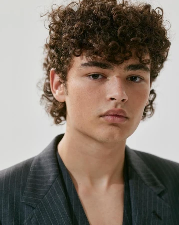 Headshot of a young adult male with curly hair, wearing a dark pinstripe blazer in a studio setting, showcasing fashion and beauty.