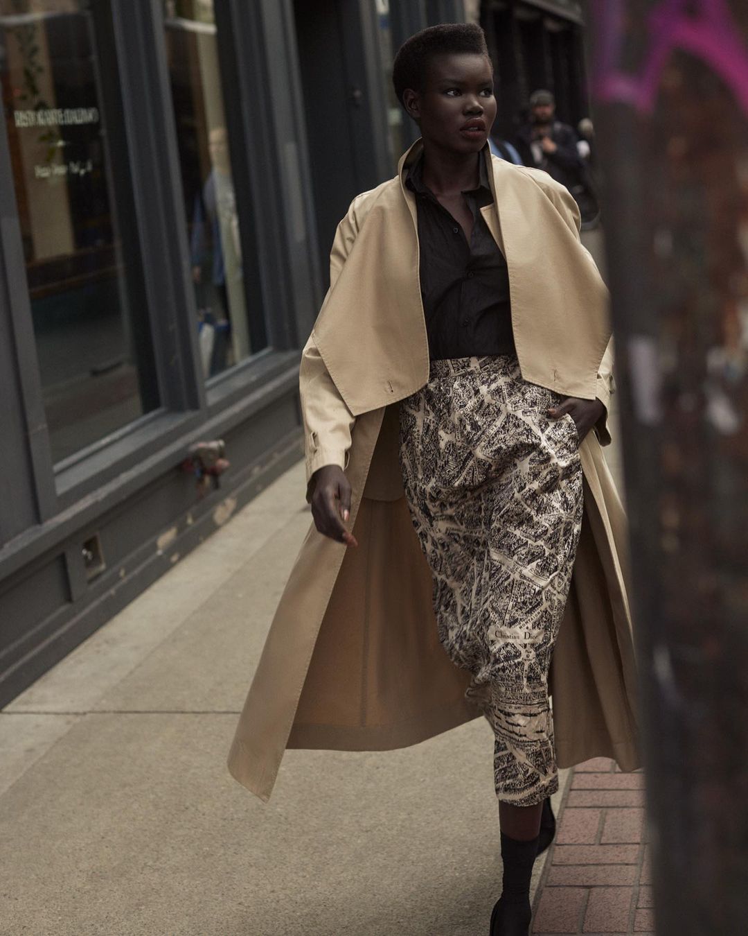 A fashionable African-descent woman strides confidently down a city sidewalk wearing a trench coat and patterned pants.