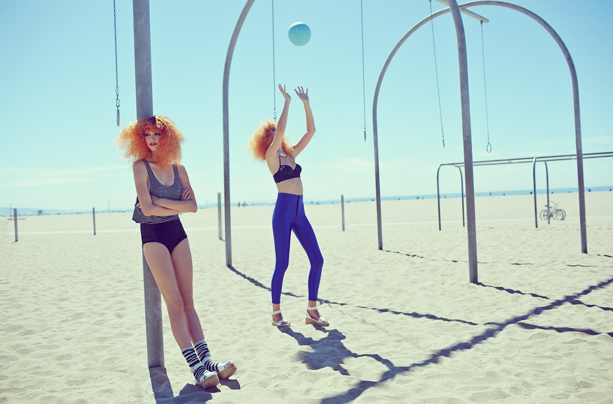 Two women with afro hairstyles are on a beach. One is leaning on a pole, the other is reaching for a blue ball. Beach and sky.