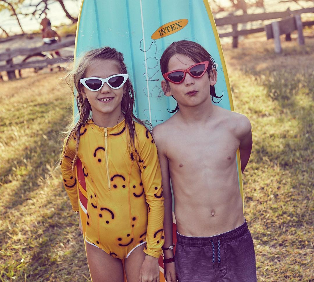 Two kids pose with surfboard at beach: girl in smiley-face swimsuit, boy in swim trunks, both wearing cat-eye sunglasses, summer vibe.