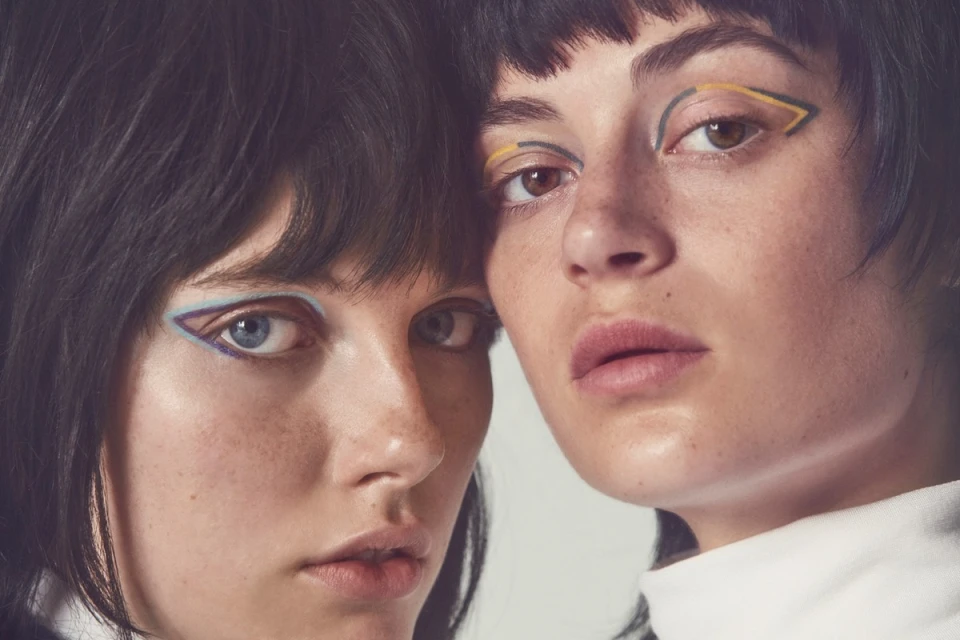 Close-up studio shot of two women with dark hair and graphic eyeliner, posing in front of a neutral background, beauty and fashion.