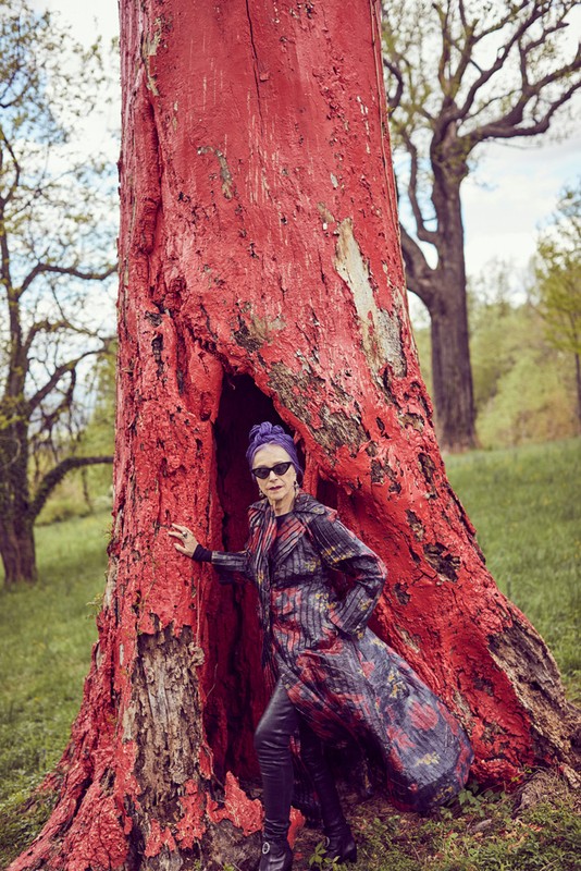 Mature woman in patterned coat standing next to a large red tree with sunglasses and a purple turban in outdoor setting.