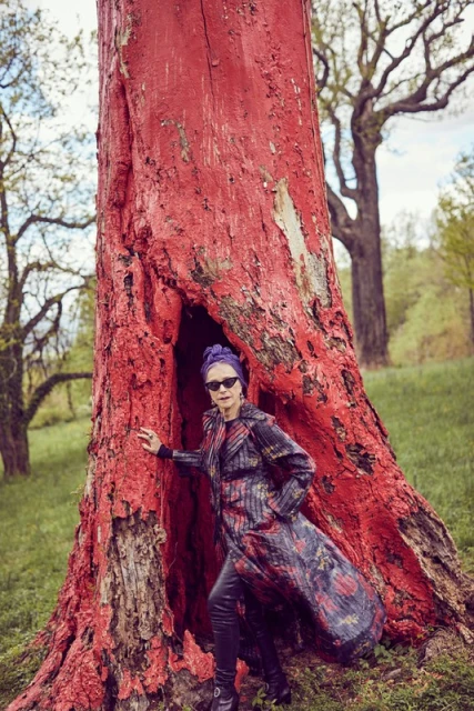Mature woman in patterned coat standing next to a large red tree with sunglasses and a purple turban in outdoor setting.