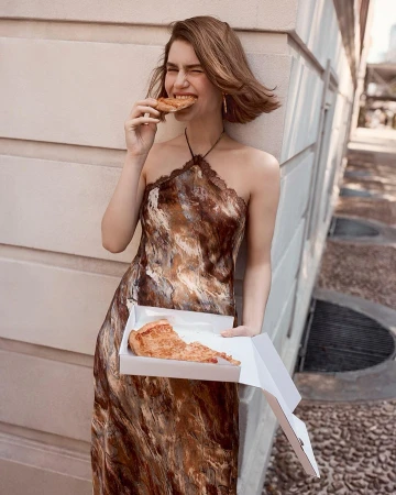Young woman in a patterned dress enjoys pizza outdoors, holding a slice to her mouth and pizza box in hand against a textured wall.