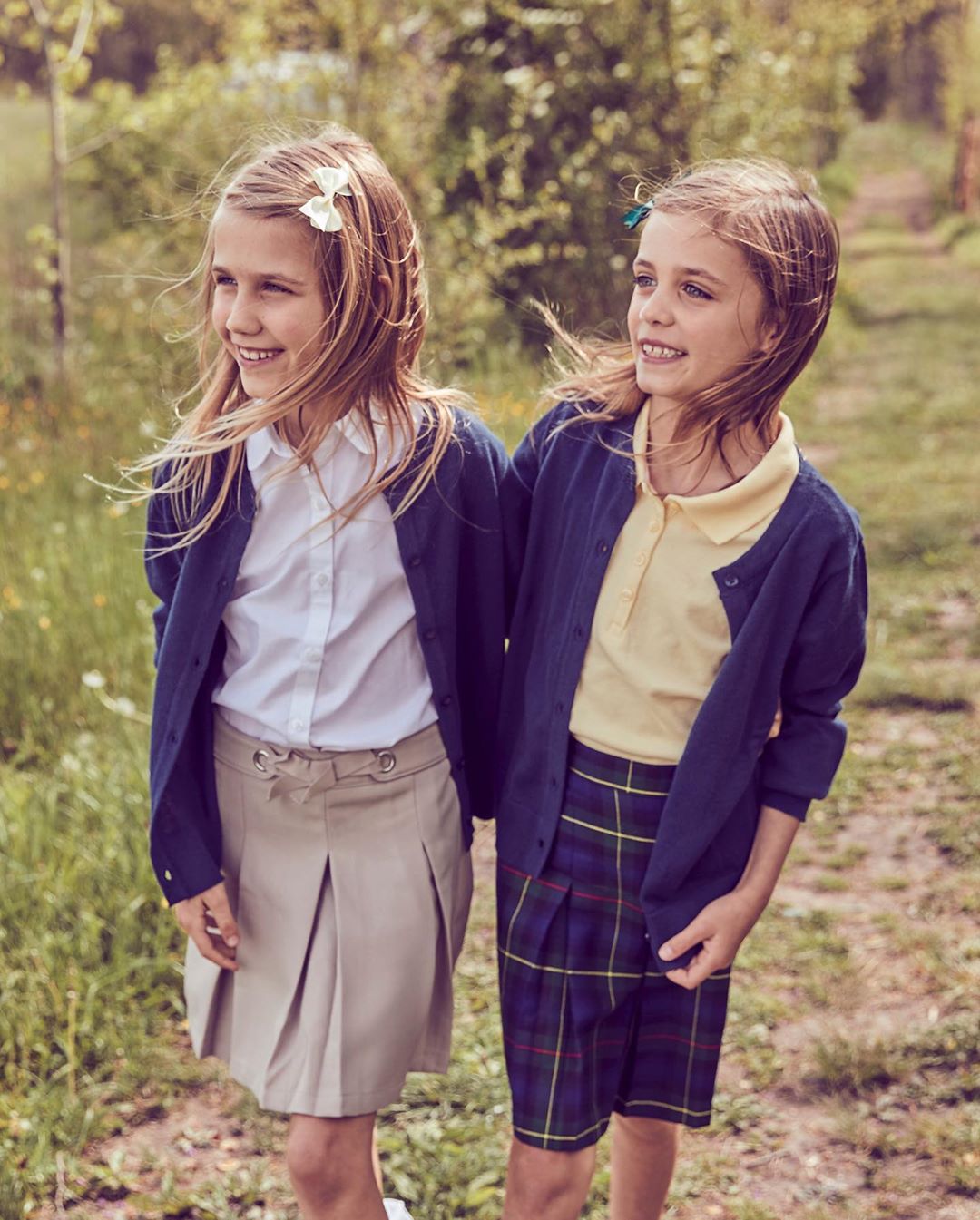 Two smiling girls in school uniforms stand outdoors surrounded by greenery. They both have blonde hair and are facing to the left.