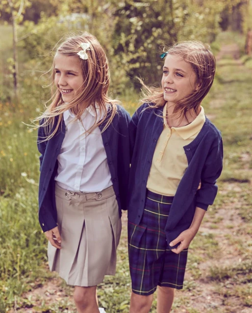 Two smiling girls in school uniforms stand outdoors surrounded by greenery. They both have blonde hair and are facing to the left.