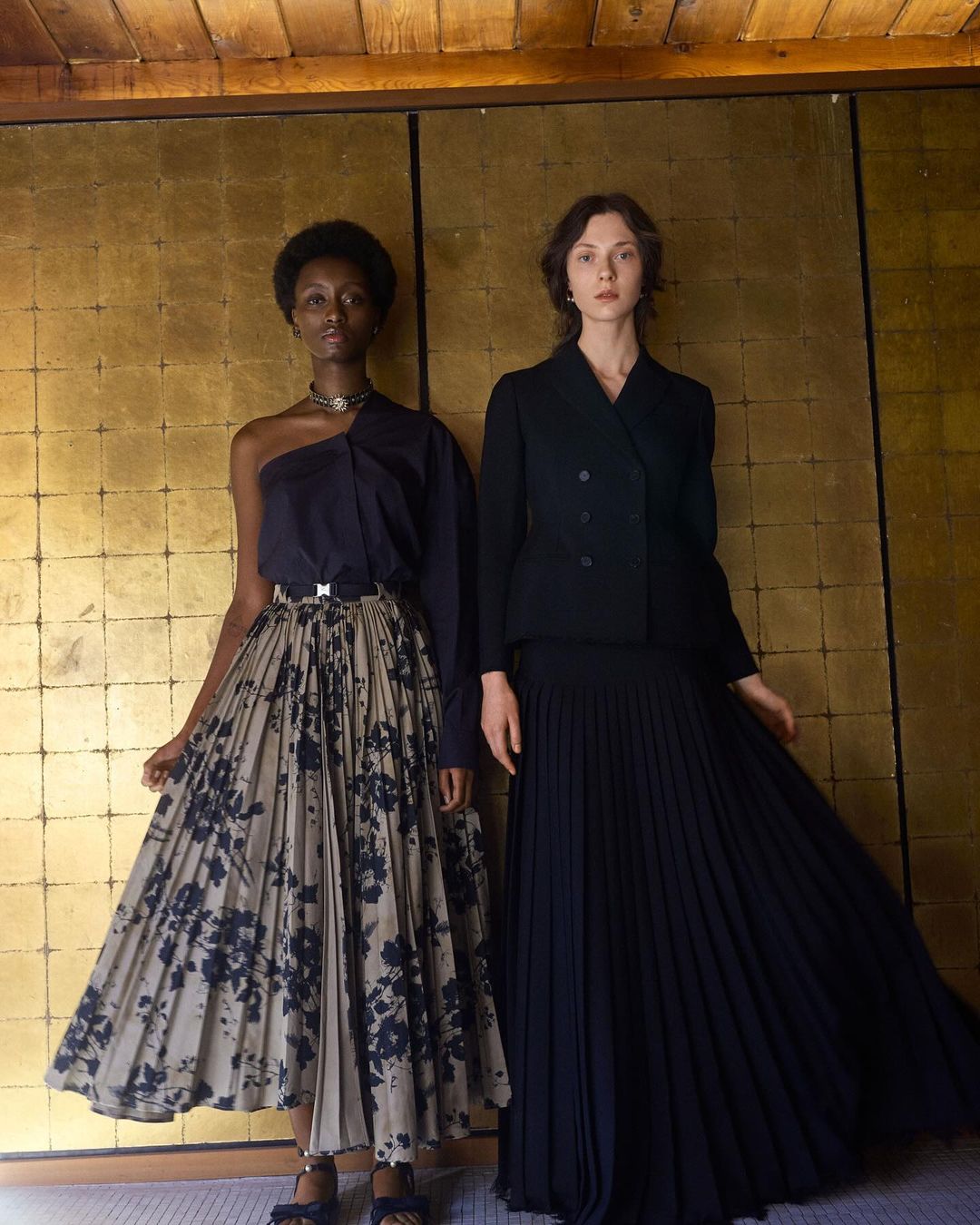 Two women in elegant dresses pose against a gold-paneled wall, showcasing a blend of modern and classic fashion elements.